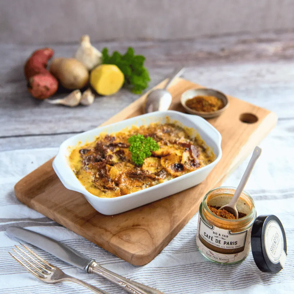 Kartoffelgratin auf Holzbrett mit Gewürzglas im Vordergrund und Gemüse im Hintergrund