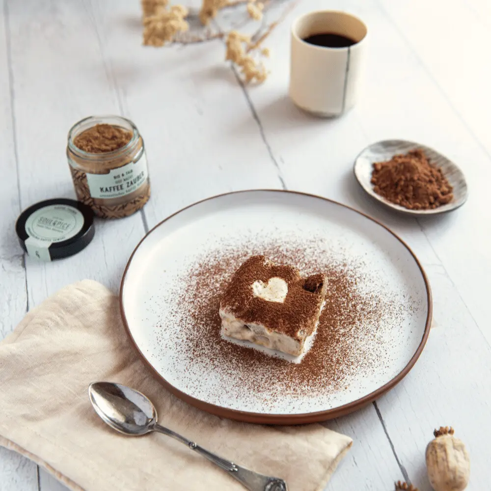 Tiramisu mit Herz auf Teller inklusive Kaffeezauber-Gewürz Glas, Kaffeetasse und Blumen